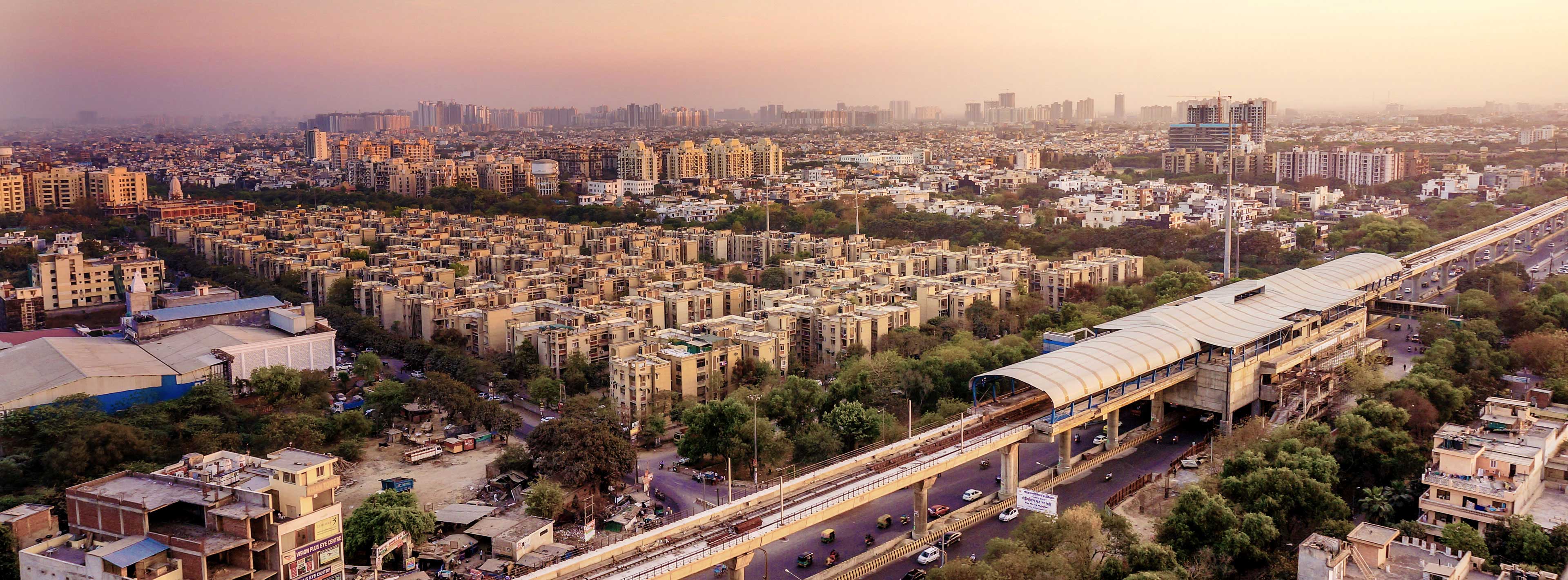 Luftaufnahme einer Stadtlandschaft mit mehreren Wohnblocks, die dicht nebeneinander stehen. Eine Straßenbahnlinie verläuft über eine Brücke, neben der mehrere Autos fahren. Im Hintergrund sind moderne Gebäude und eine leicht neblige Skyline sichtbar.