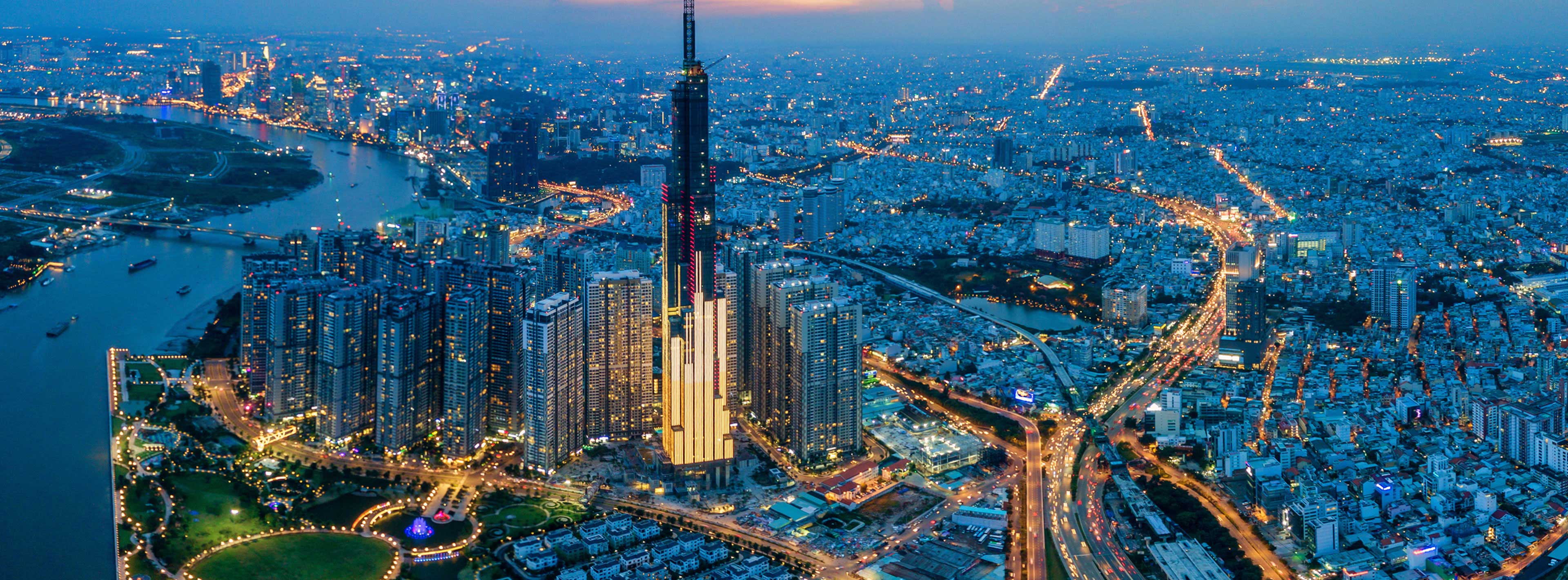 Eine Panoramaaufnahme von Ho-Chi-Minh-Stadt bei Dämmerung. Im Vordergrund sind moderne Wolkenkratzer, darunter der Landmark 81, sichtbar. Die Stadt erstrahlt in bunten Lichtern, während der Saigon-Fluss im Hintergrund verläuft. Die Szene vermittelt ein Gefühl von urbaner Lebhaftigkeit.