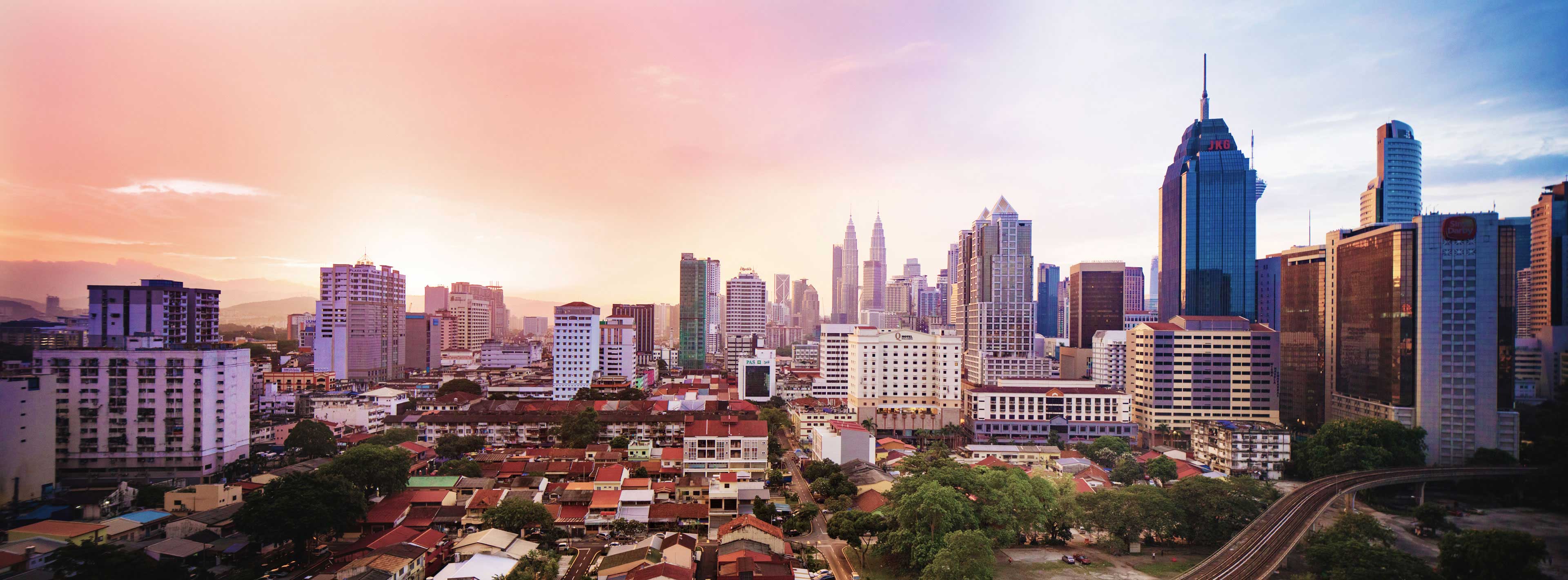 Eine Stadtansicht von Kuala Lumpur bei Sonnenuntergang. Hohe Wolkenkratzer, darunter die berühmten Petronas Towers, sind sichtbar. Im Vordergrund sind flachere Gebäude und Wohnhäuser mit roten Dächern zu sehen, umgeben von Bäumen und Straßen. Der Himmel zeigt sanfte Farben in Pastelltönen.