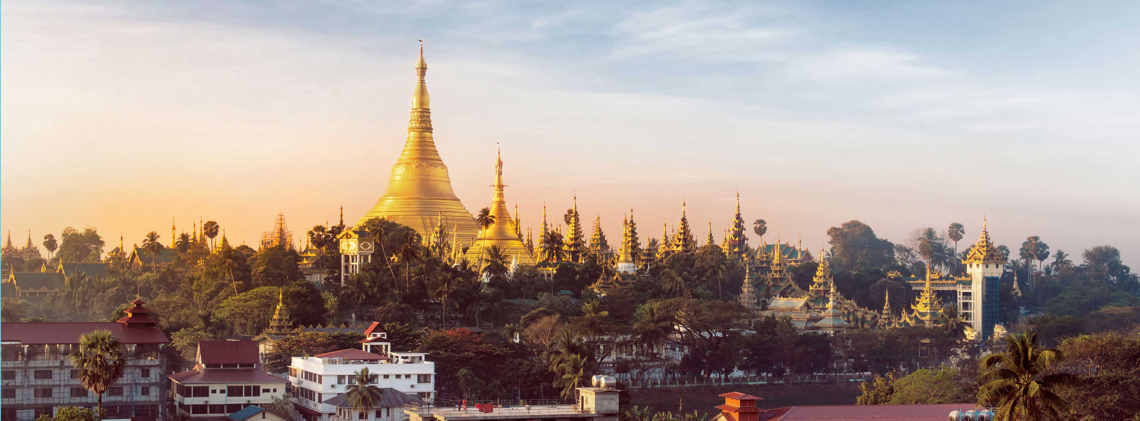 Die Aufnahme zeigt die goldene Stupa der Shwedagon-Pagode in Yangon, umgeben von zahlreichen weiteren Pagoden und grünen Bäumen. Im Hintergrund erhellt das sanfte Licht des Sonnenaufgangs die Szene. Die Architektur hebt die kulturelle Bedeutung und Schönheit des Ortes hervor.