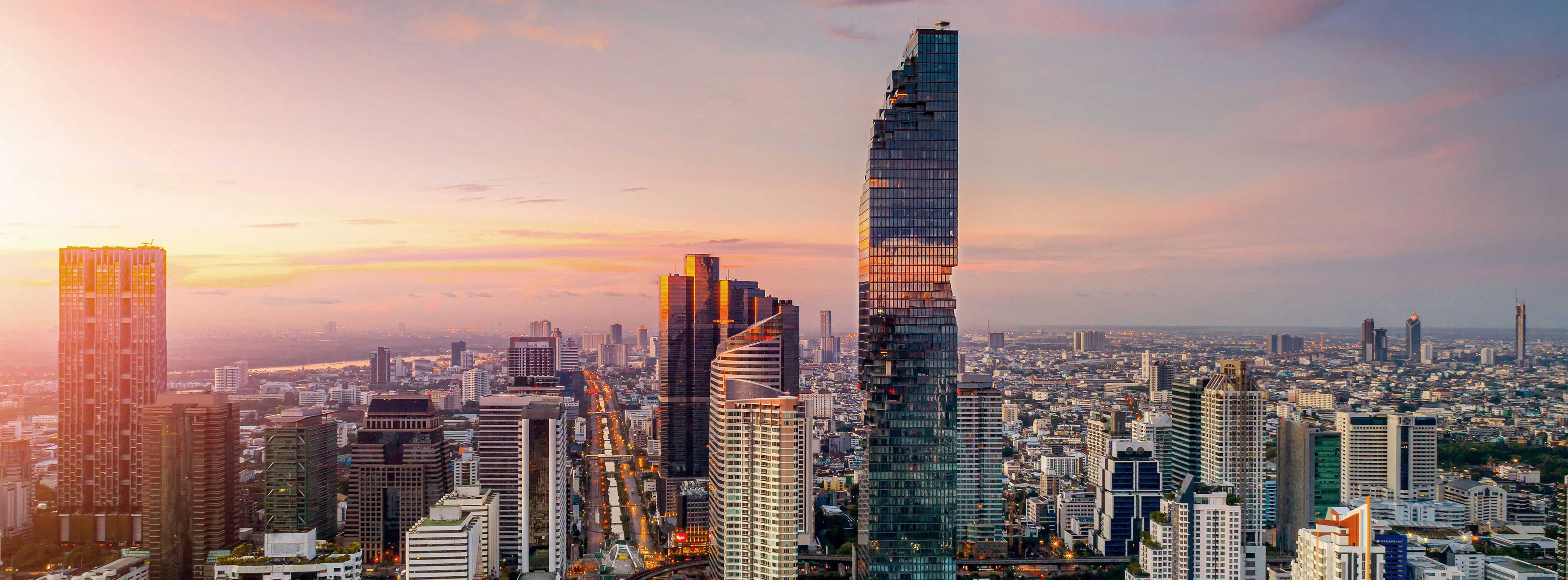 Eine weite Stadtansicht zeigt beeindruckende Wolkenkratzer unter einem farbenfrohen Sonnenuntergang. Im Vordergrund sind moderne Gebäude sichtbar, während sich im Hintergrund die Stadtlandschaft erstreckt. Die Himmelstöne variieren von Gelb bis Rosa, was eine ruhige Atmosphäre schafft.