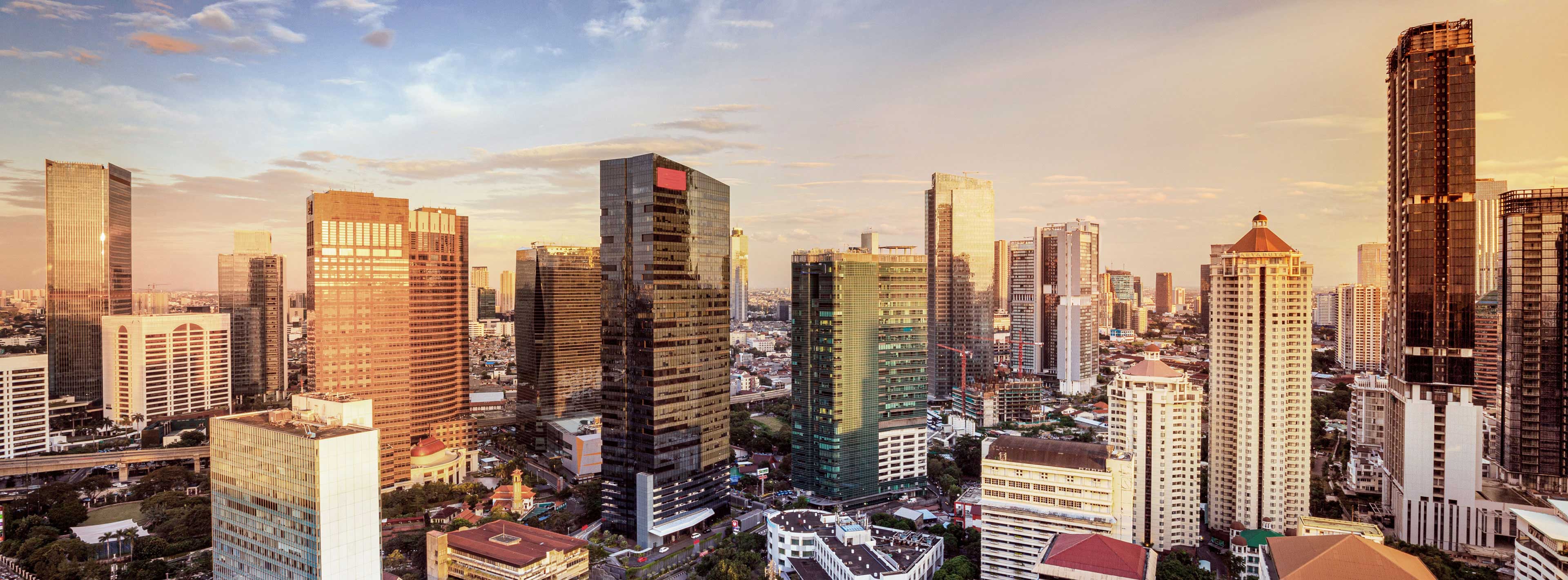 Eine weite Panoramaansicht einer modernen Stadtlandschaft zur Dämmerung. Hochhäuser mit Glasfassaden spiegeln die warmen Farben des Sonnenuntergangs wider, während sich darunter Straßen und kleinere Gebäude erstrecken. Die Szene vermittelt ein Gefühl von urbaner Dynamik und Zukunft.